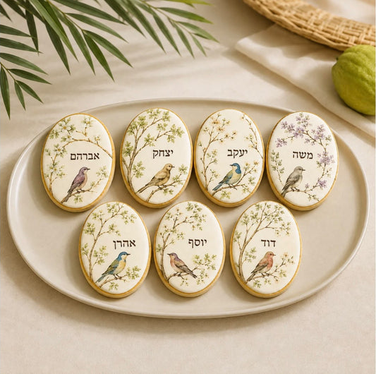 Seven oval sugar cookies decorated with bird illustrations and Hebrew text, arranged on a light beige ceramic plate with a neutral background and soft decorative shadows.